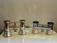 Photo showing two pairs of opera binoculars side by side on a reflective surface, one gold colored and one silver colored, both with mother of pearl inlay.