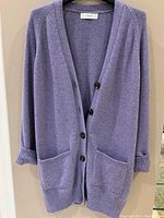 Front view of lavender cashmere cardigan with button closure and front pockets hanging on a hanger against a white wall.