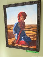 Framed oil painting of a man in a red turban and blue clothing sitting on desert sand dunes, viewed from the front.