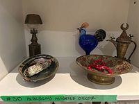 Overall shot of metal lamp, ceramic dish with sage, glass bird, marble sphere, brass ewer, and pedestal bowl with red beads on white shelf