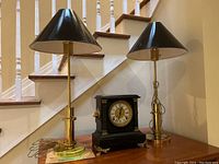 Front view showing both brass lamps with black shades and the black mantel clock on a wooden table.