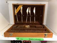 Open wooden case containing 5 knives secured vertically on the inside lid and 12 pieces of forks and spoons arranged in velvet-lined compartments inside the case.