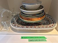 Stack of assorted ceramic bowls, plates, platters and clear acrylic plate holders on shelf.