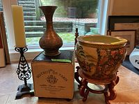 Photo showing the ceramic pot with colorful design on wooden stand next to tall metal vase and candle holder with candle, with footstool in front