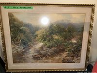 Frontal image of large framed print showing man fishing in a river, with trees and rocks surrounding him. Frame is gold-tone with a wide beige mat board.
