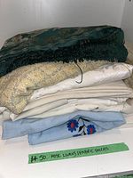 Stack of assorted linens and fabric pieces, including a dark green silky embroidered fabric, yellow textured textile, plain beige and cream linens, and a light blue floral embroidered piece.