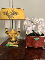 Yellow vintage-style table lamp with floral pattern and yellow fabric shade next to faux wooden planter with white artificial flowers on a decorative wooden table.