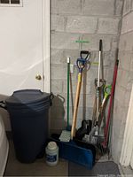 Wide view of lot showing trash bin, various yard tools including shovels, rakes, mop, broom, and ice melt container against concrete block wall
