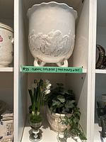 Photo of white ceramic pot with scalloped rim and leaf design on four feet, placed on a shelf with artificial plants and small artificial flowers underneath.
