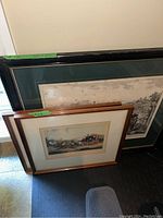 Three framed prints leaning against a wall, largest with black frame and green matting, smaller prints with wooden frames