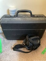 Black hard plastic carrying case with handle and shoulder strap camera bag placed on floor in front of a light wall