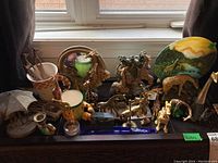 Wide photo showing multiple giraffe-themed items including mugs, figurines, plate, candle holder, mirror with giraffe figures, and small animal figurines all arranged on a black surface in front of window.