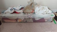 Wide view of assortment of folded table linens and lace pieces on and in top of clear plastic container placed against wall on carpet floor.