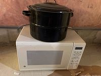 Photo showing black enamel stock pot with lid placed on top of a white GE microwave model JE114OWNC 001 on a basement floor.