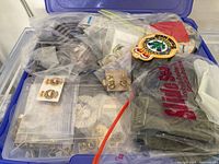Plastic bin filled with multiple bags containing patches and pins, including a visible red and green embroidered patch and a 'Slite Seal' branded cloth item.