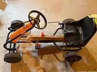 Side-angle photo of the orange and black pedal go-kart showing the frame, steering wheel, and one pedal missing.