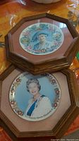 Two framed collector plates on a table covered with plastic; one depicts Queen Elizabeth in formal attire, the other Queen Mother in blue hat and floral decor.