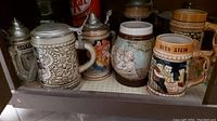 Shelf showing a variety of beer steins including floral relief, painted scenes, and traditional styles.