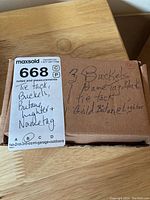 Photo of closed box with handwritten label describing contents: 3 buckles, 1 name tag (black), 1 tie tack, gold butane lighter