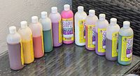 11 bottles of Simply Tempera paint in assorted colors, partially used, placed in a row on a glass table.