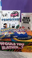 Photo of four games: Would You Rather? party game green box, Man Bites Dog card game small box styled as a newspaper, DOS and Skip-Bo card games.