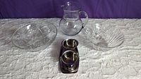 Glass pitcher, large cut glass bowl, medium lead crystal bowl, and MCM stainless steel sugar, creamer, and tray set on white quilted surface with purple background