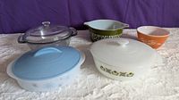 All five kitchenware pieces on display: three covered casseroles and two Pyrex bowls, arranged on a white surface with purple background.