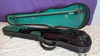 Black violin inside an open hard case with teal interior lining, resting on a white surface under purple background