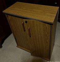 Front angled view of rustic wood cabinet showing woodgrain finish, two doors with wooden handles, and flat top surface.