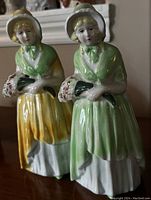 Two enamel painted female figurines in bonnet hats and dresses, one green and yellow, one green and white, holding small bouquets.