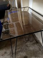 Photo of a rectangular clear glass top table with black metal frame and legs against a wall and concrete floor showing scratches on the glass surface.