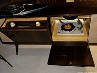 Photo of 1960s stereo console with open fold-down doors showing radio tuner with two knobs on left and turntable with 45 RPM record on right side.