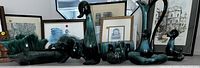 Seven ceramic and glass vases including green, cobalt blue, and brown items displayed on a table with framed art prints in background