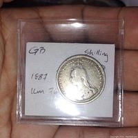Obverse side showing Queen Victoria portrait and inscription, in plastic flip holder with handwritten note reading 'GB 1887 Shilling'