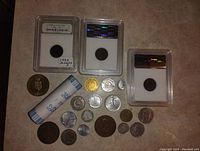 Photo showing three certified coins in plastic holders, one labeled 1935 Arizona, sealed roll of nickels, and various loose US and world coins spread out on surface.