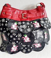 Front view of handbag showing black scalloped panels with white snowflakes and snowmen patterns, and red leather top with buckle detail.