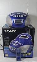 Front view of the Sony CFD-E75 showing sapphire blue unit with integrated speakers, front LCD display, control buttons, telescoping antenna, sitting on top of original product box. Remote control placed in front.