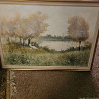 Framed landscape painting showing two figures by a lake under trees with a village in the background.