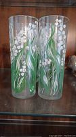 Two tall clear glasses with hand-painted green leaves and white Lily of the Valley flowers, placed side by side on a shelf.