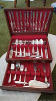 Silverware set in open wooden case showing knives, forks, spoons, and serving pieces neatly arranged in velvet-lined compartments.