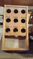 Top view of rectangular wooden bean bag gameboard with nine holes labeled with different point values, showing aged wood with chips and scratches.