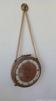 View of antique round wall clock hanging by decorative rope showing wooden face, Roman numerals, glass cover, and metal frame.