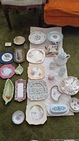 Top-down view of 25 pieces of assorted vintage china and dishes arranged on carpet showing variety of shapes, colors, and designs including floral and bird motifs.