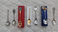 Seven assorted collector souvenir spoons on light quilted background; includes various crest designs and different metal tones.