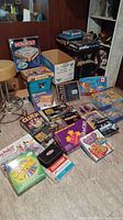 Photo of multiple boxed board games including Monopoly, Guess Who?, Clue Jr, and several other titles stacked on and around a rug.