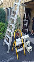 Photo showing large 7.5 ft aluminum step ladder, yellow metal step stool, and small wooden step stool in front of brick wall.