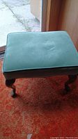 Footstool standing on a reddish carpet, showing its green upholstery and wooden legs.