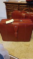 Two reddish-brown Samsonite suitcases on carpet in front of wooden dresser.
