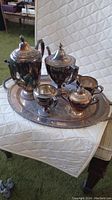 Full view of six-piece silver tea service set on a silver tray placed on a white quilted surface.