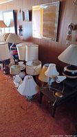 Wide view of 14 assorted table lamps and shades placed on carpet. Lamps vary in base style and shade design, many shades are stained and discolored.